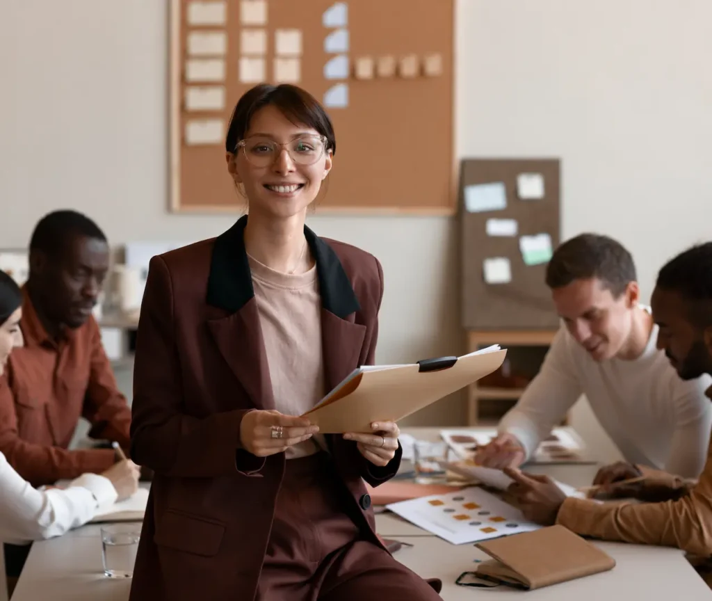 A smiling HR leader holding a folder while her team collaborates in the background, illustrating what human resources are and how C2 Essentials helps you understand the heart of your business.