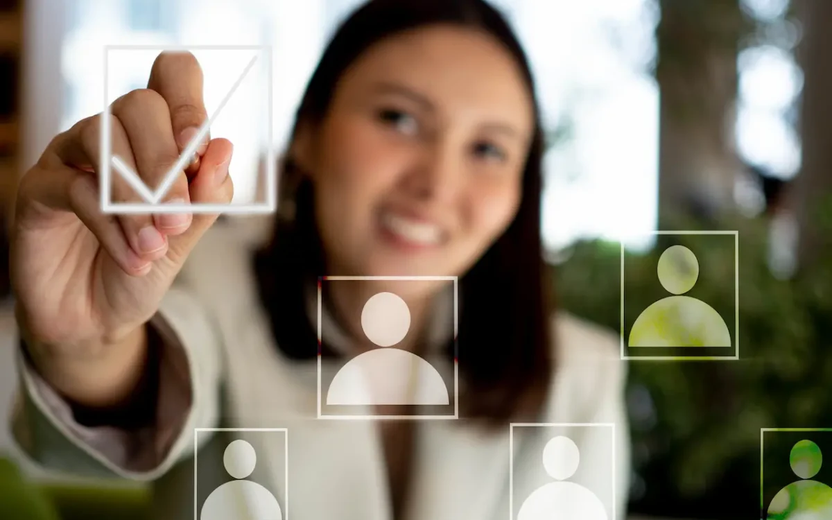 A close-up view of a person's hand with a pen, checking a virtual checkbox over a translucent person profile icon, representing Human Resources decoding success through precise selection and evaluation for your business with C2 Essentials.